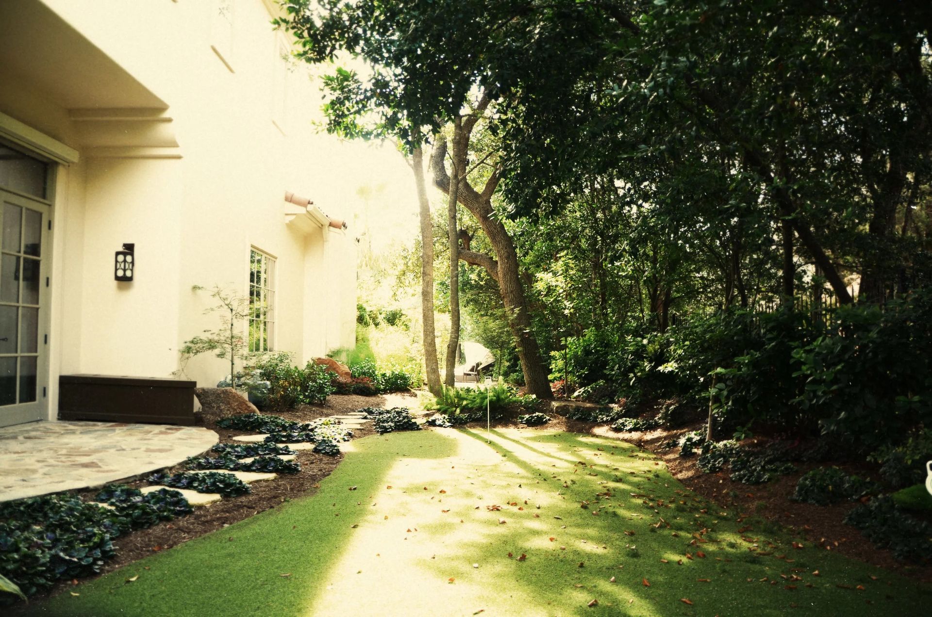 A sunlit backyard garden with manicured grass, garden beds, and mature trees next to a cream-colored home.