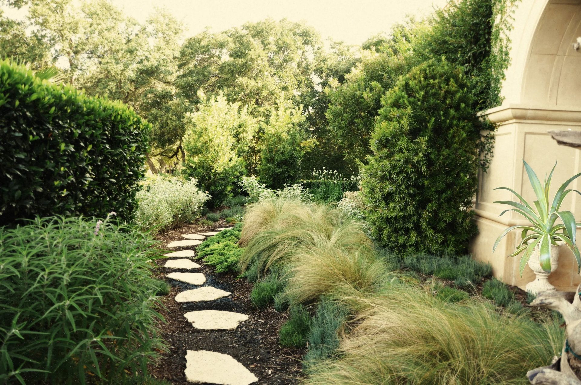 A stone stepping-stone path winds through a lush, green garden toward a stone archway in the distance.