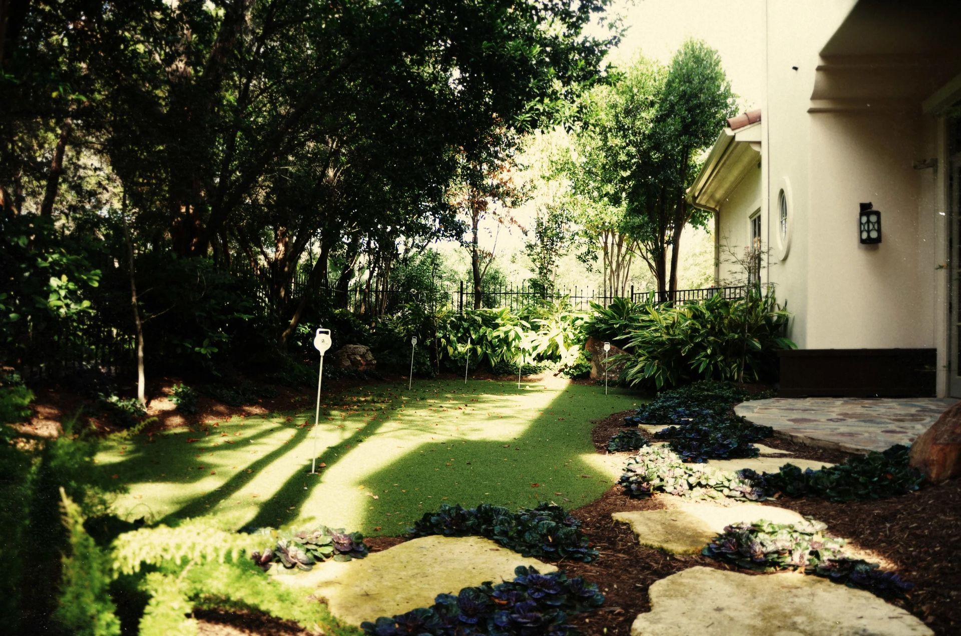 A landscaped backyard with a putting green, stone stepping stones, lush trees, and a light-colored house exterior.