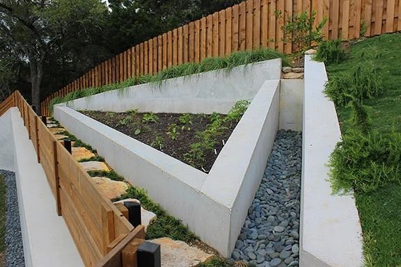 Triangular raised garden bed, concrete construction, with plants, set against a wooden fence on a hillside.