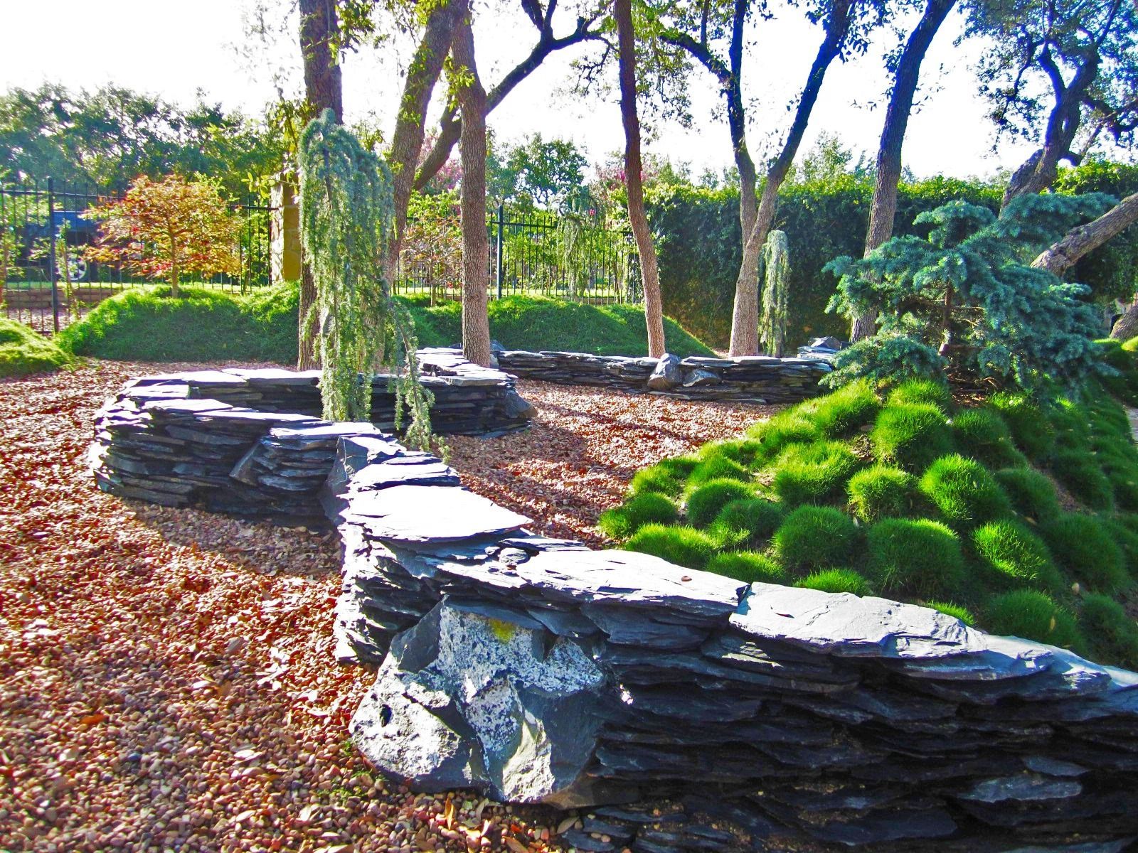 A winding stacked slate wall borders a garden bed with mounds of green foliage and trees in a wood-chipped landscape.
