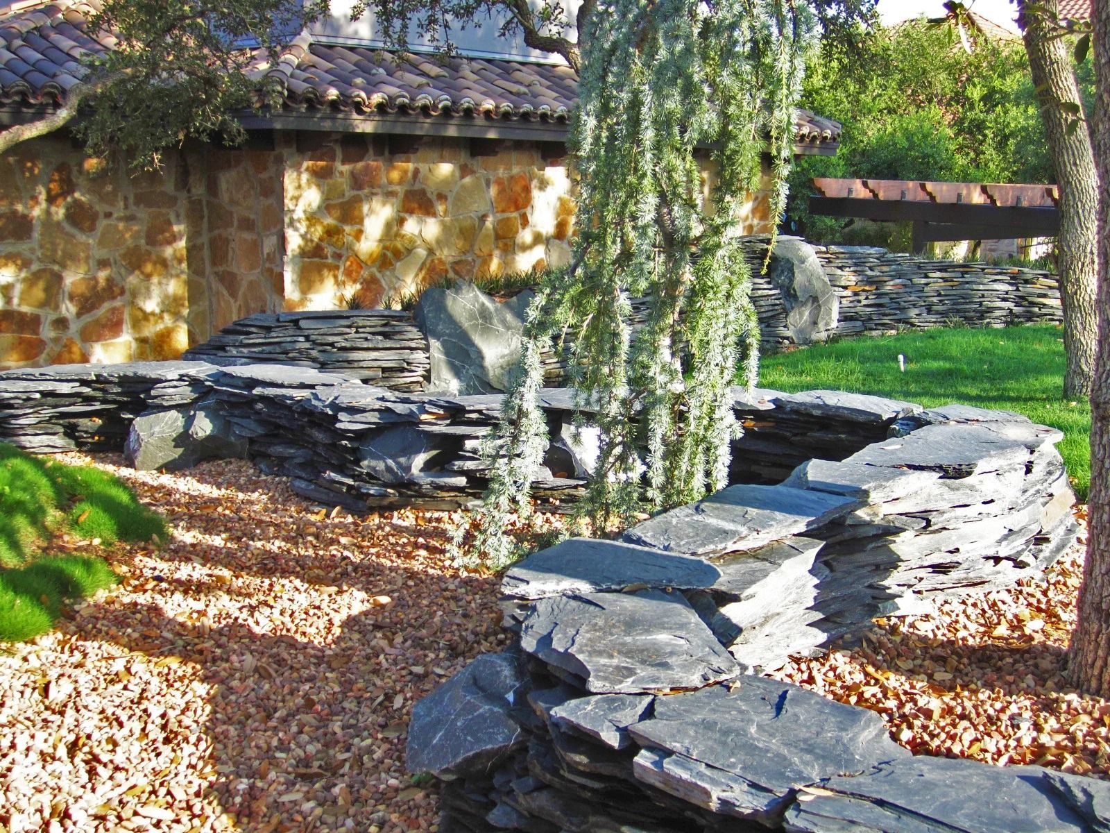 A curved stacked slate wall winds through a garden bed filled with brown mulch, near a stone house and green grass.