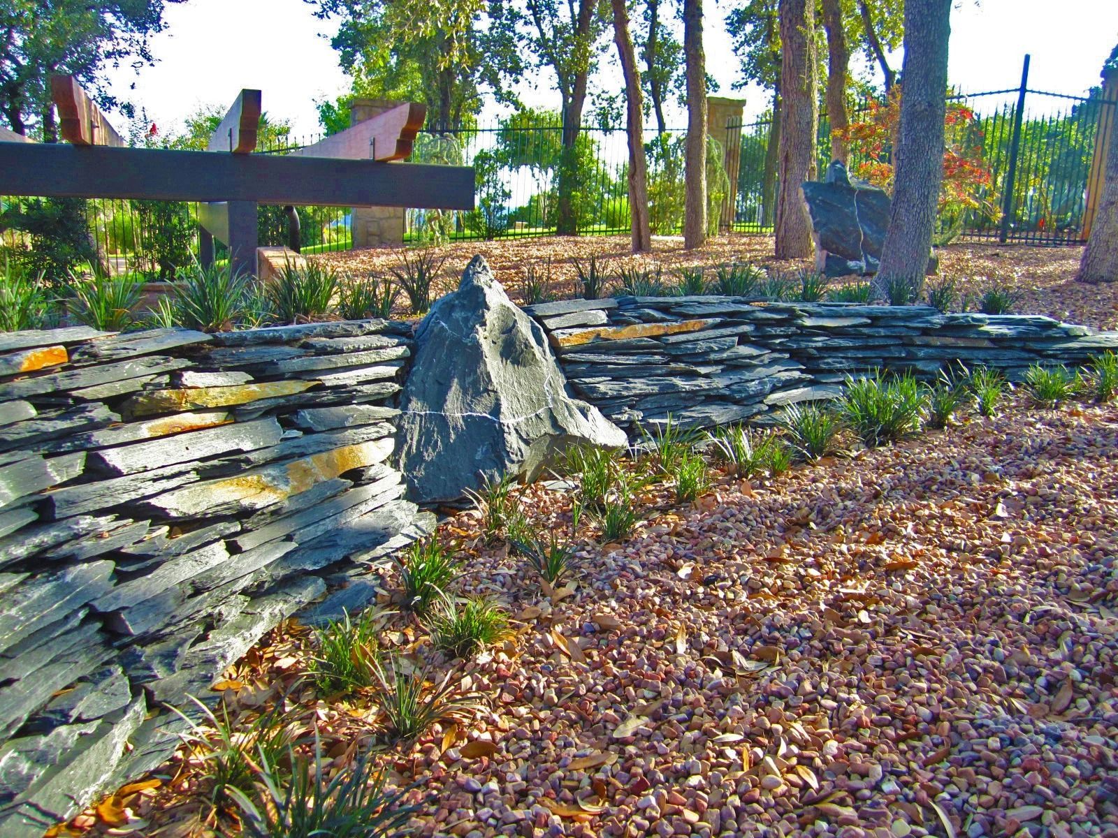A landscaped garden features a dark, stacked slate retaining wall accented by a large, upright boulder and small plants.