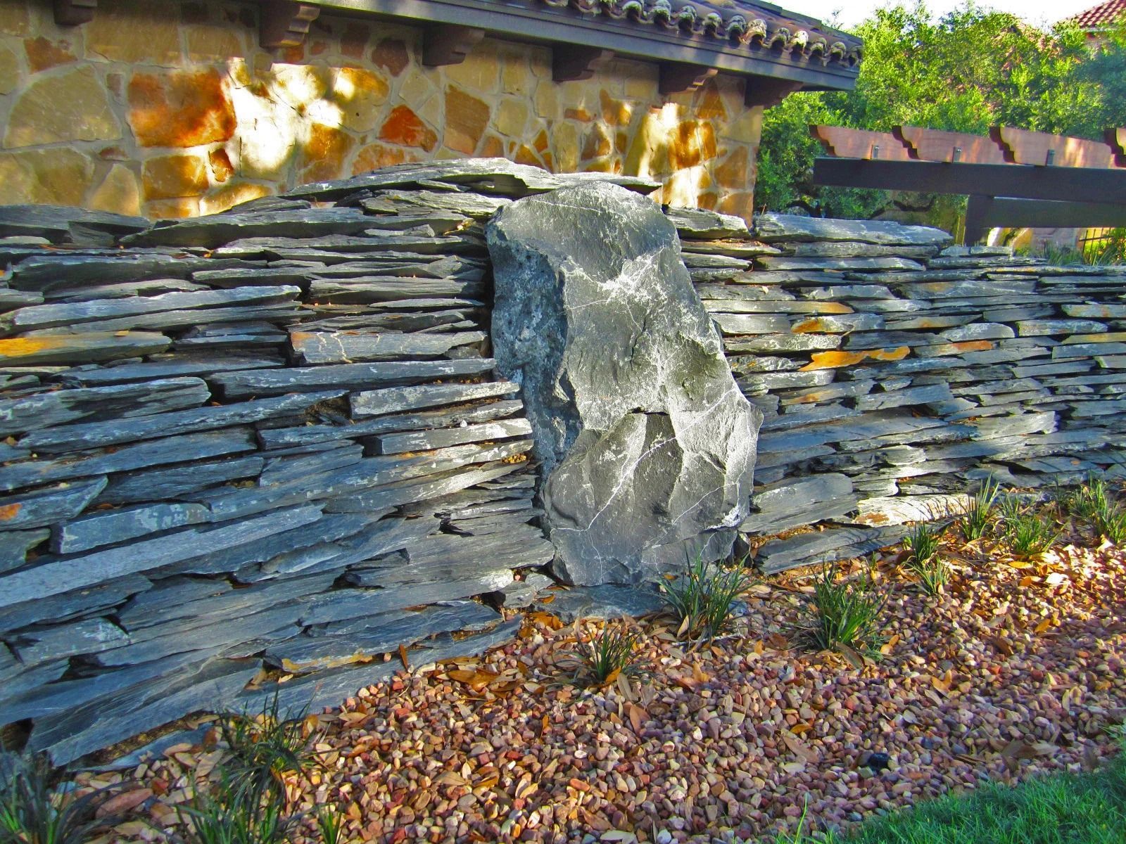 A decorative garden wall of stacked grey slate featuring a large, vertical natural boulder centered in the stone masonry.
