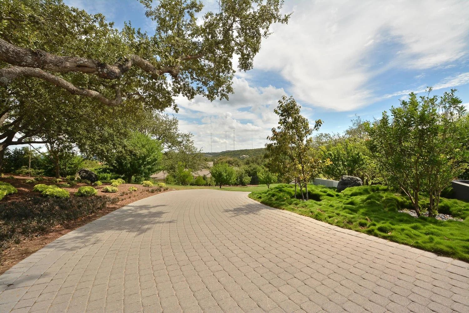 Brick driveway through a landscaped yard, leading towards trees and a sunny sky.
