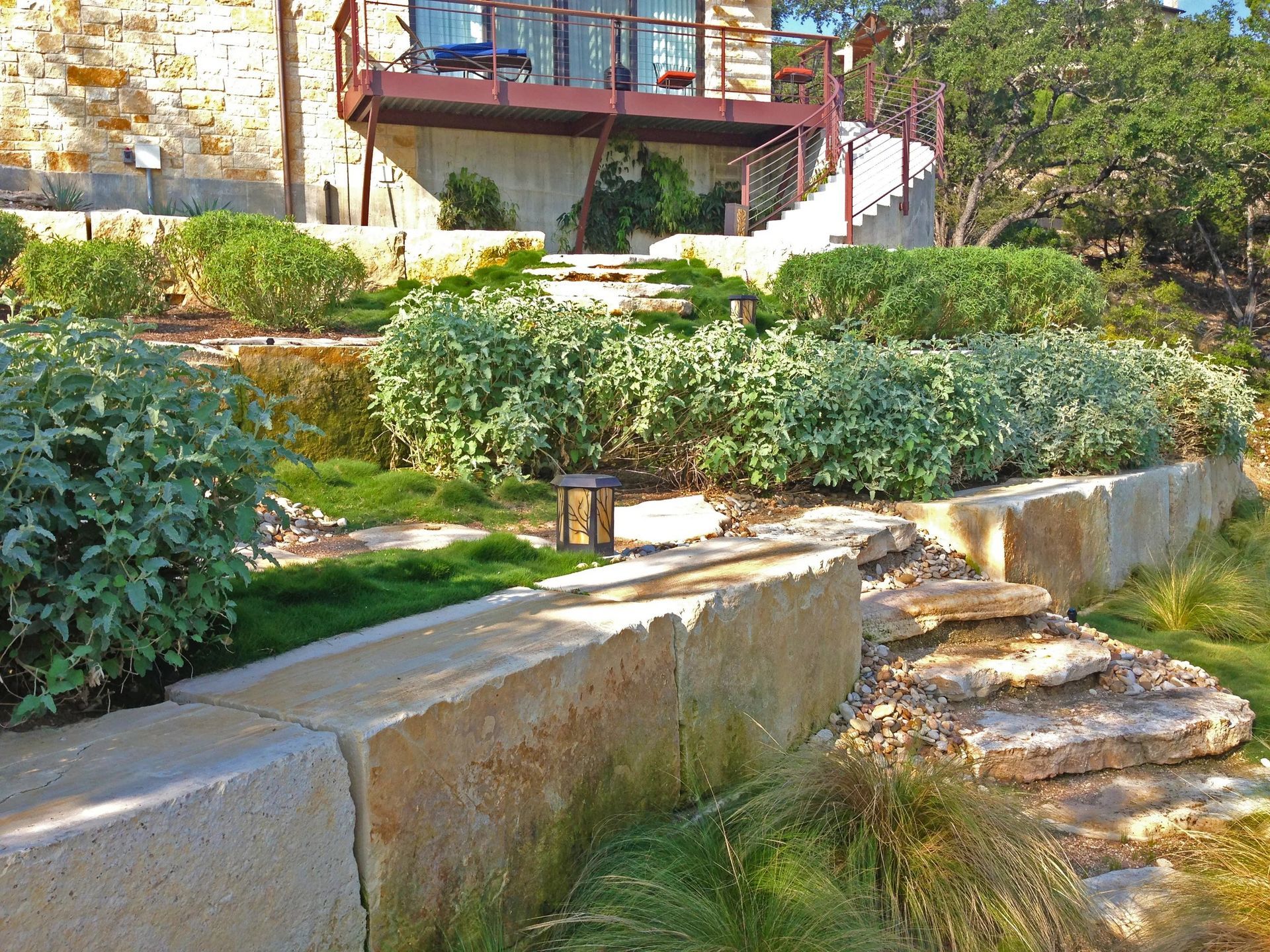 A tiered stone retaining wall landscape leading to a multi-level deck and stone house facade under bright sunlight.