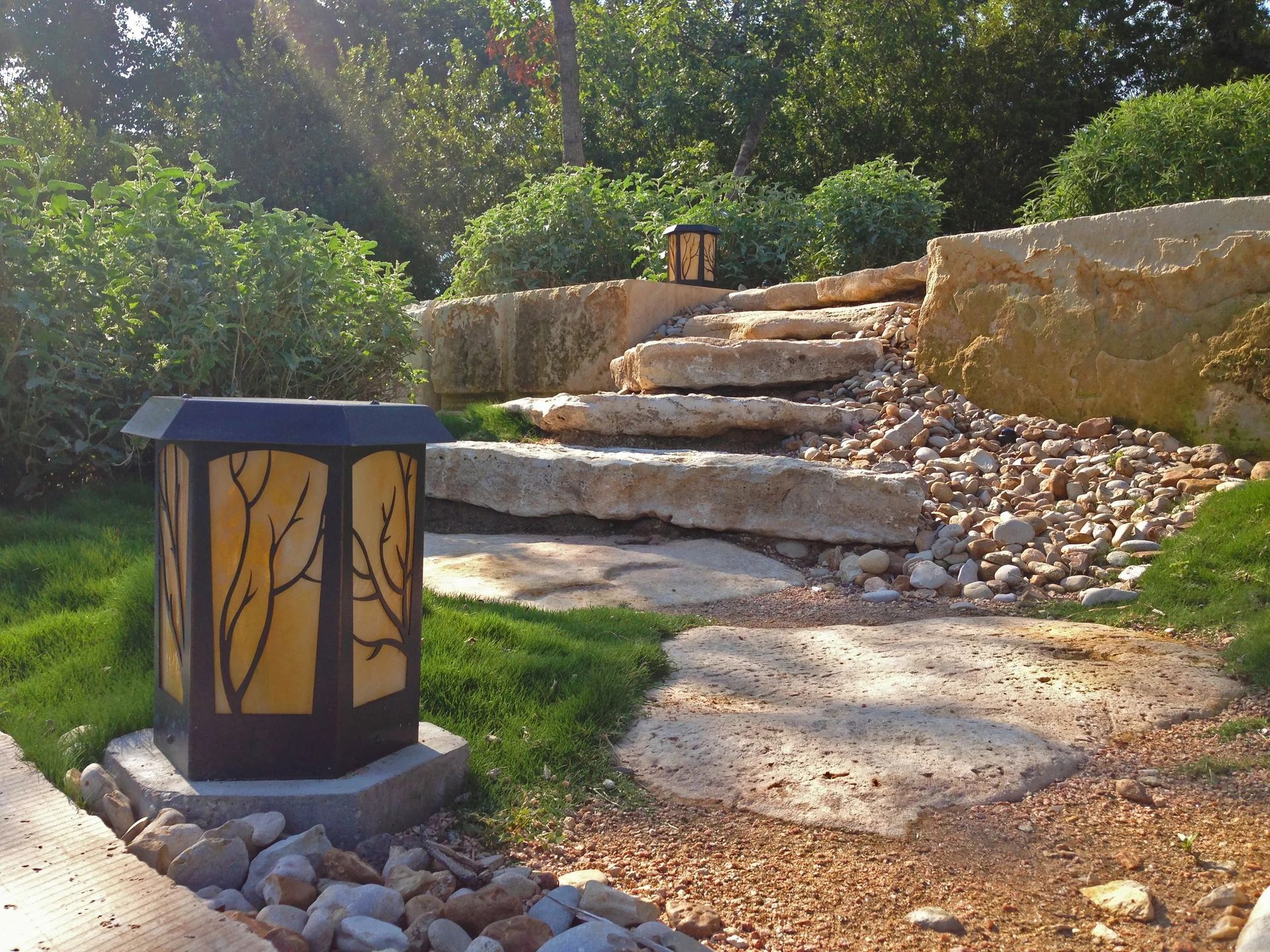 A decorative garden lamp with a branch silhouette sits on a stone pathway leading to tiered stone steps in a sunny garden.