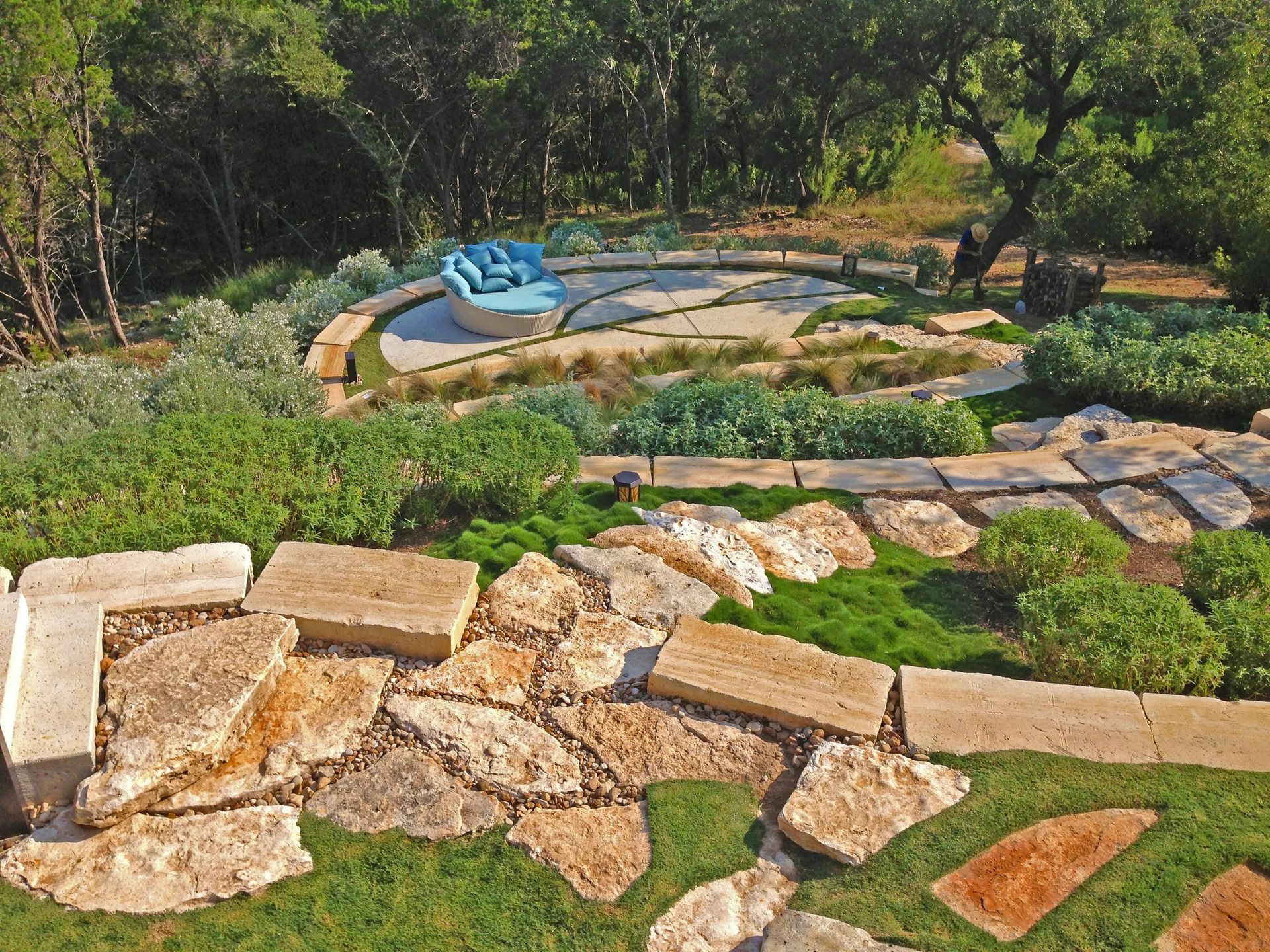 A tiered outdoor landscape with natural stone pathways, lush greenery, and a semi-circular patio featuring a blue sofa.