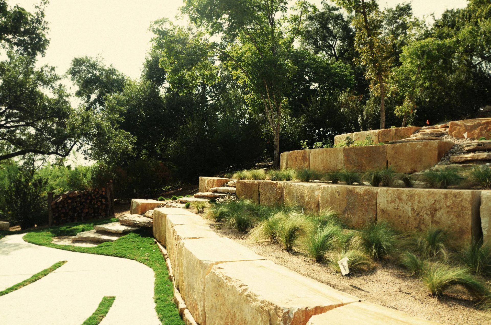 A tiered stone retaining wall landscape with grassy paths, native plants, and mature trees under a bright sky.
