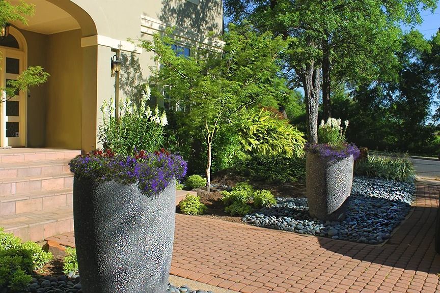 Two large textured gray planters with purple flowers flank a brick walkway leading to a house entrance.