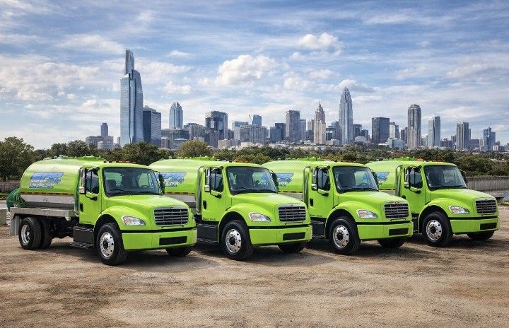 Four lime-green utility trucks parked on dirt with a city skyline in the background