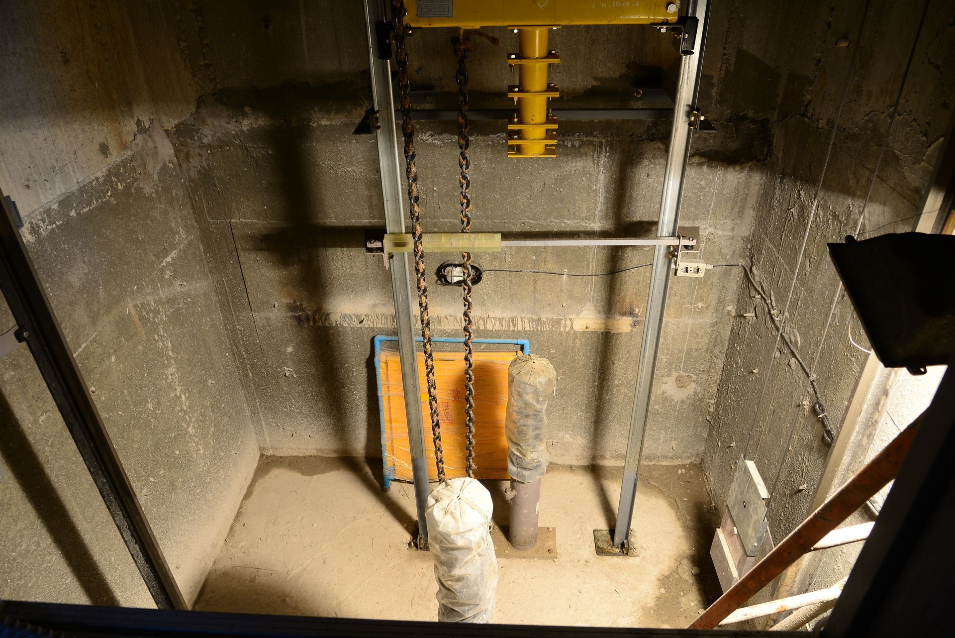 Industrial shaft with metal pipes, a yellow mechanism overhead, and a white duct in a concrete pit.