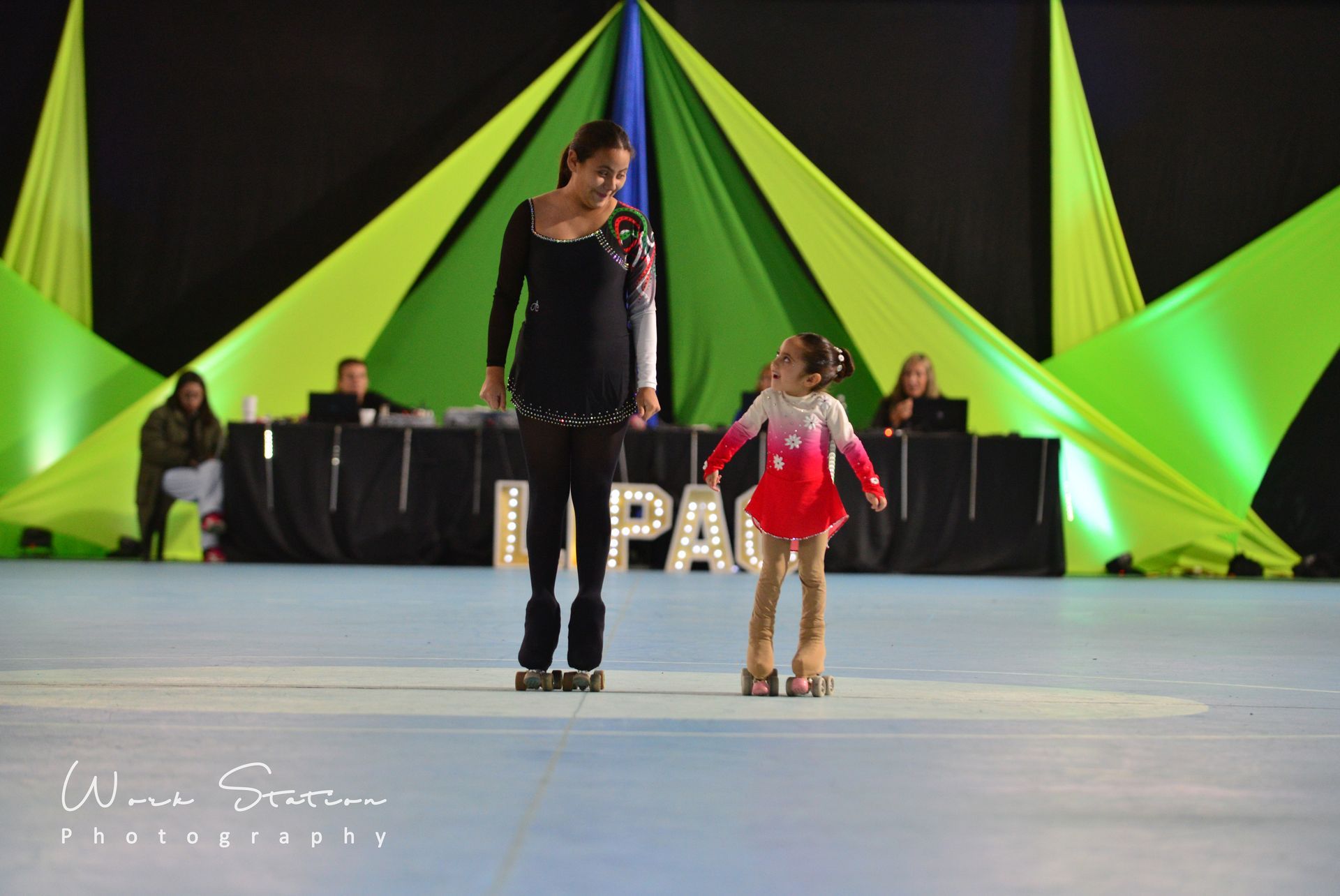 Dos patinadores se encuentran sobre una pista, uno frente al otro, frente a un escenario decorado con telas verdes y azules.