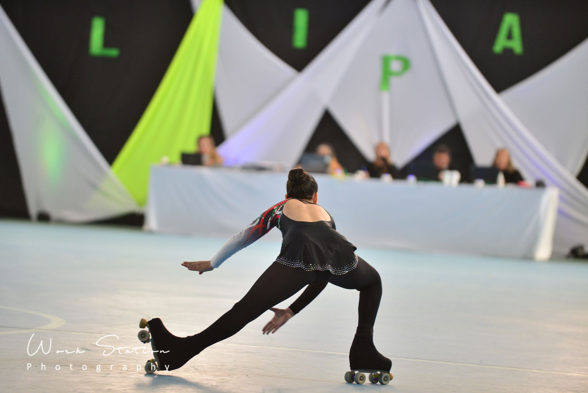 Una patinadora con un traje negro realiza una estocada profunda en una pista cubierta, con los jueces sentados al fondo.