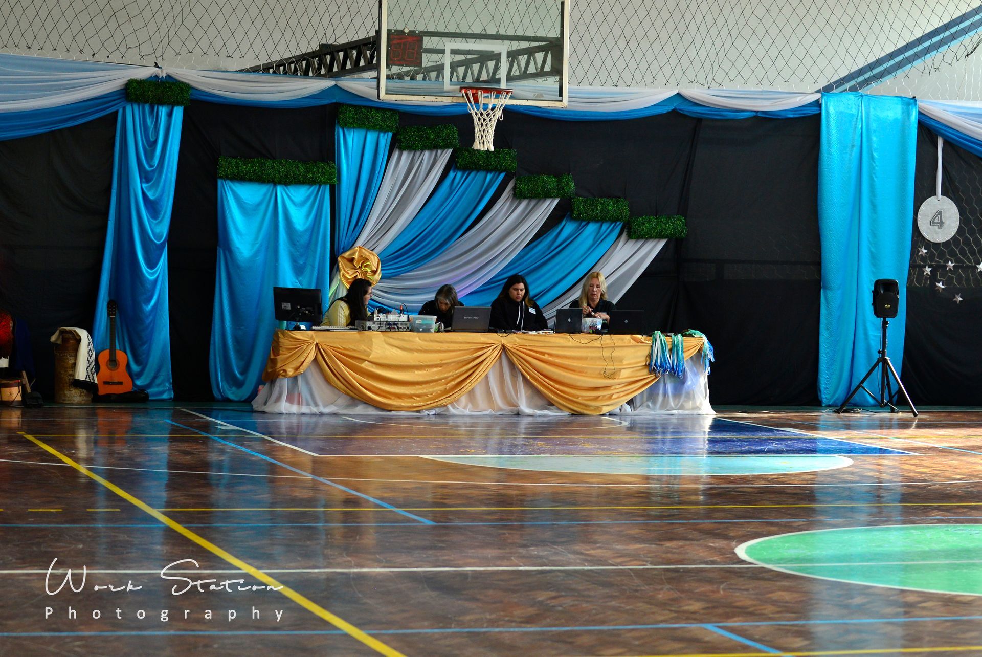 Tres personas están sentadas en una mesa cubierta con tela dorada frente a un fondo decorativo azul y blanco en un gimnasio.
