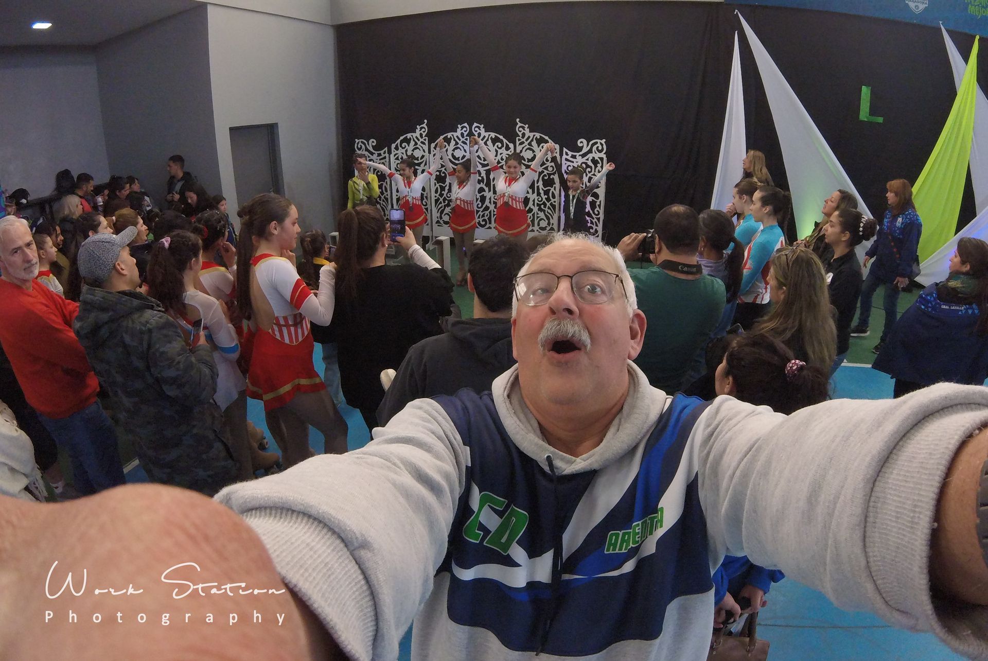 Una selfie de un hombre sonriente con bigote en un estadio deportivo, con animadoras actuando al fondo.