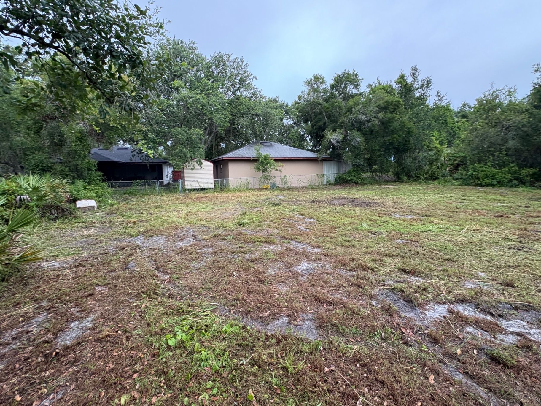 Cleared grassy lot with muddy foreground, trees, and a small house in the background under a cloudy sky
