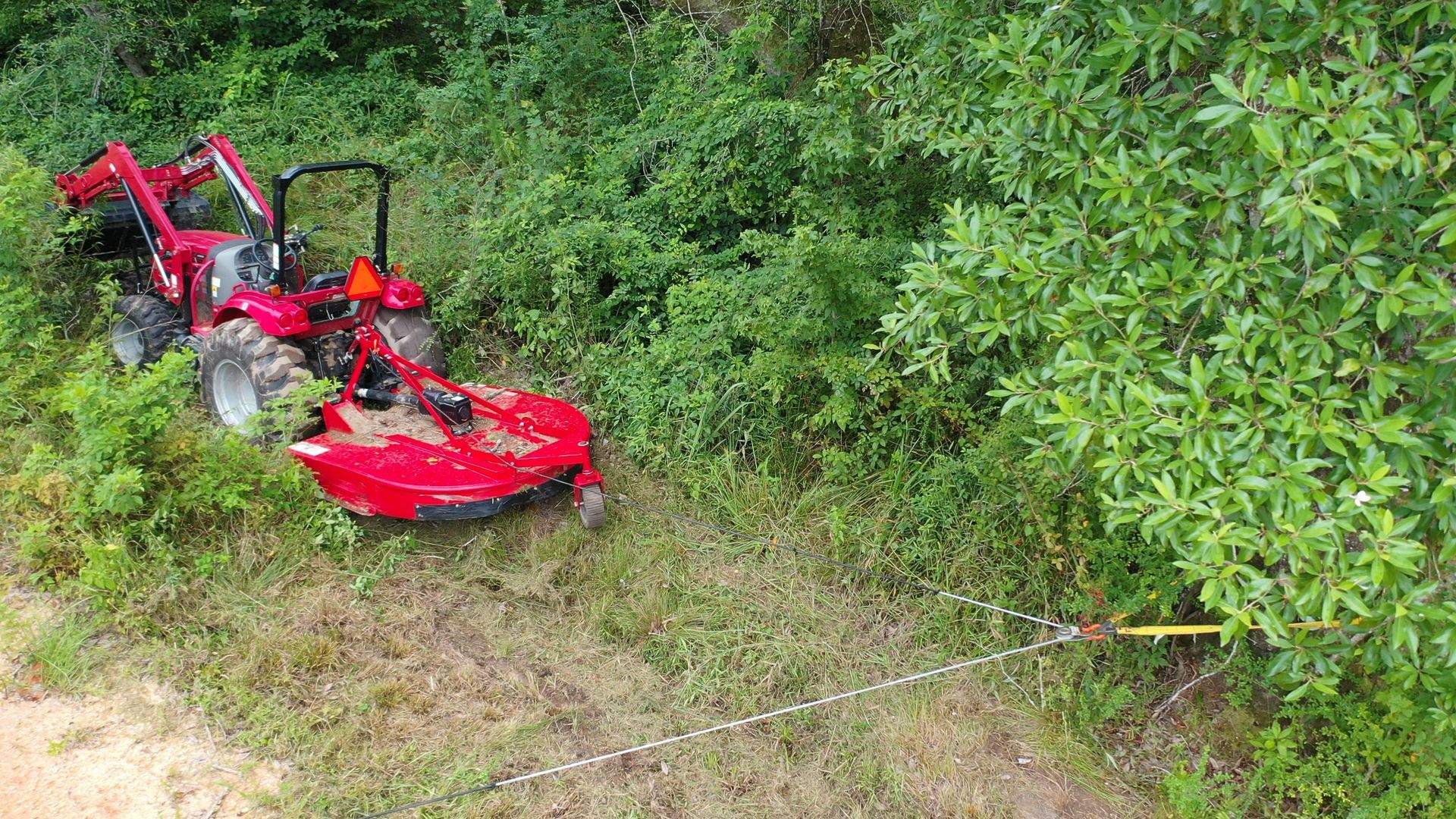 Red lawn tractor tipped over in dense green brush on a grassy slope