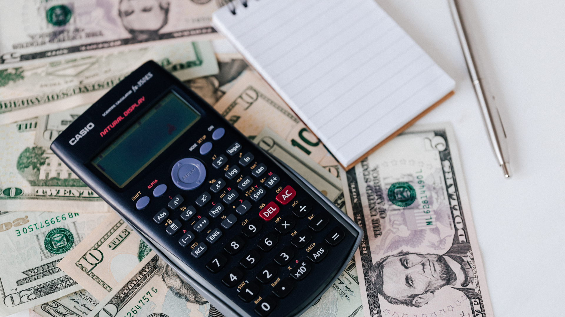 Calculator on U.S. dollar bills beside a notepad and pen on a desk