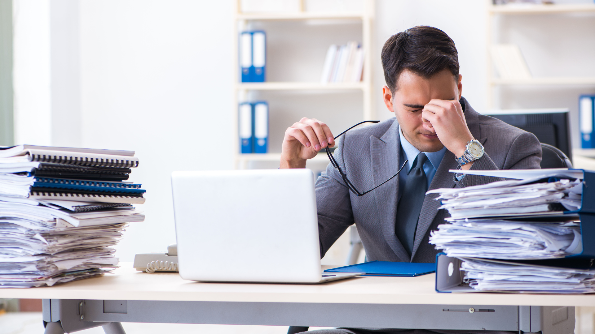 Stressed office worker rubbing eyes at desk with laptop and tall stacks of paperwork