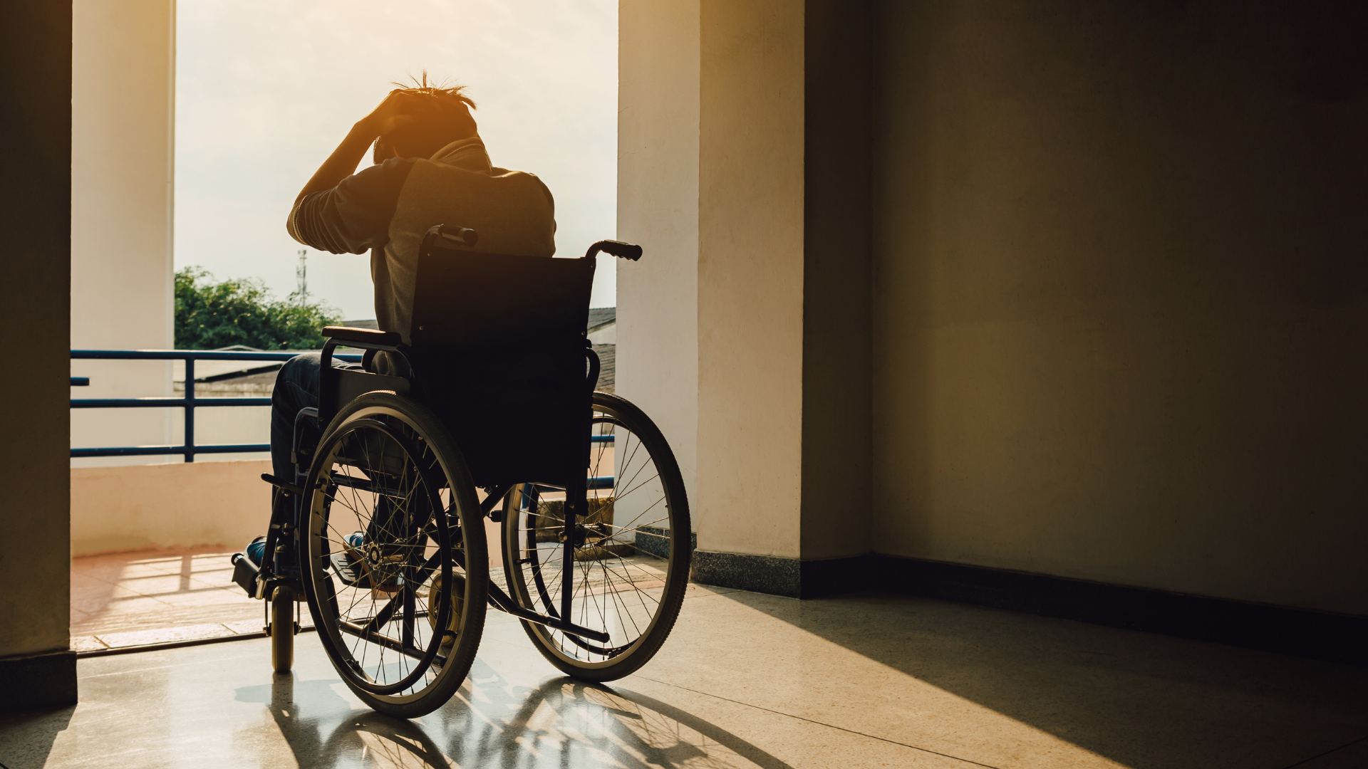 Person in a wheelchair silhouetted by sunlight in a hallway, looking out a window