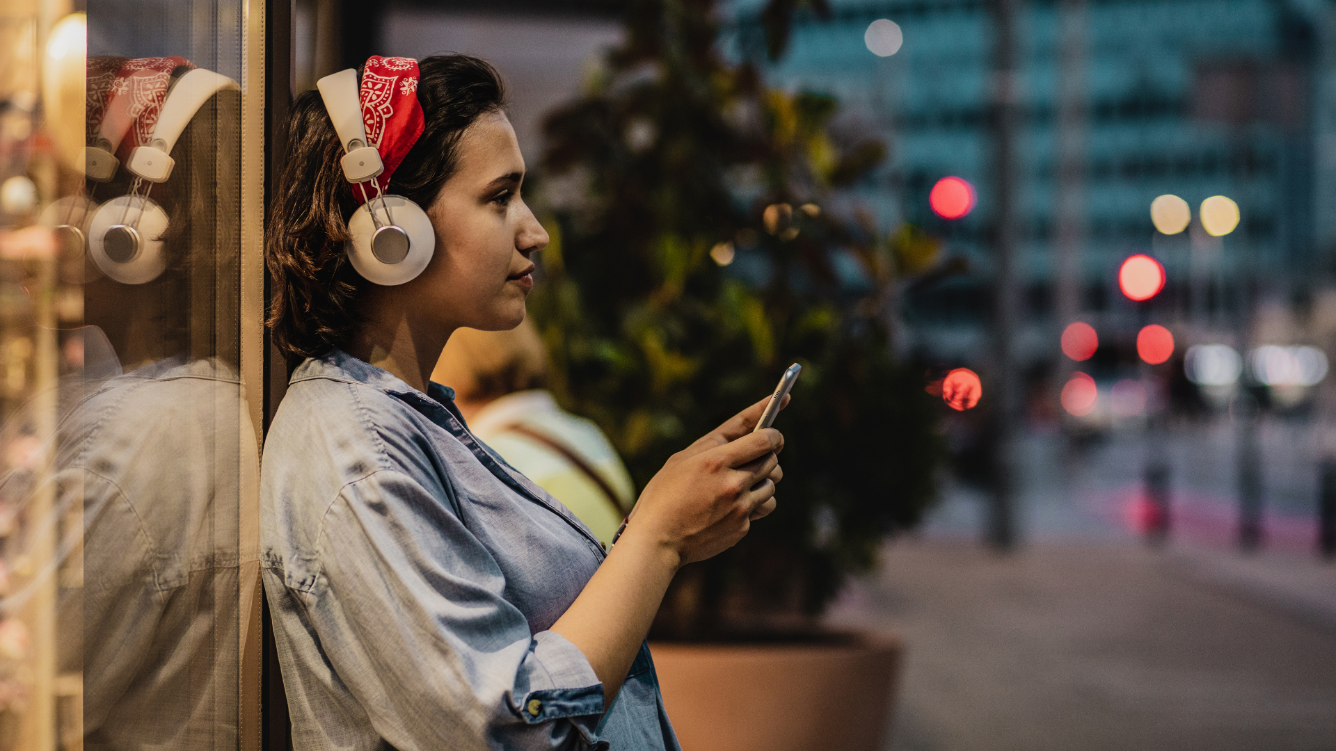 Woman wearing headphones leans by a window, looking at her phone on a city street at dusk.