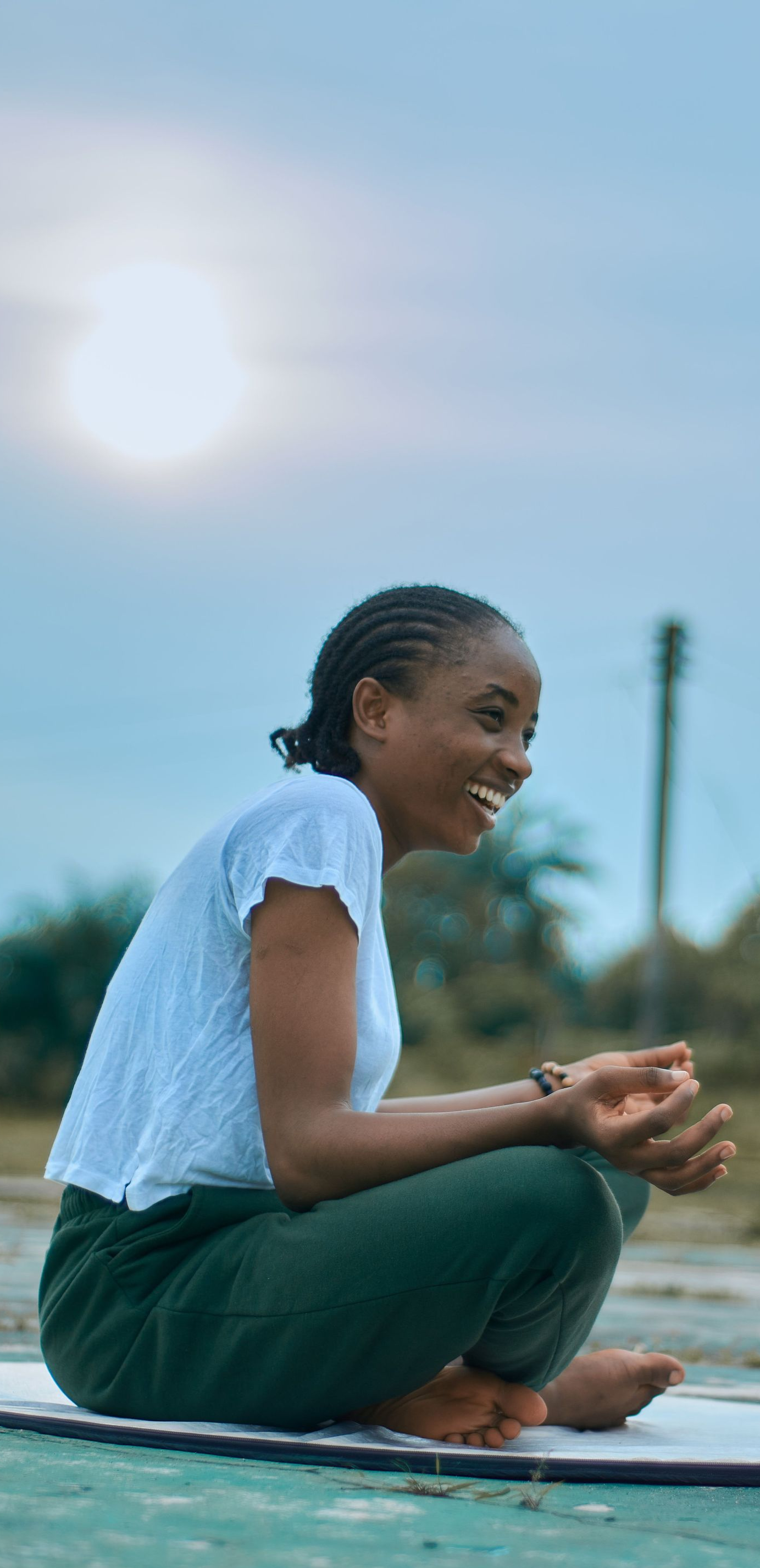 Person sitting cross-legged outdoors on a yoga mat at dusk, smiling and clasping hands.