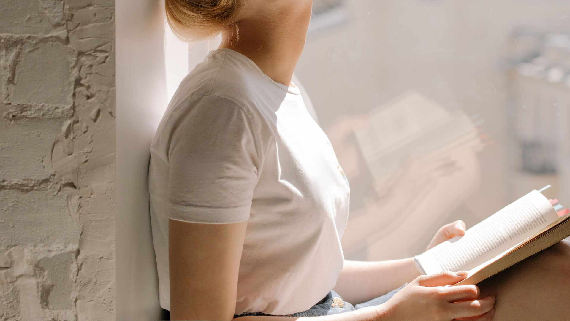 Person reading a book by a sunlit window, holding it open beside a brick wall.
