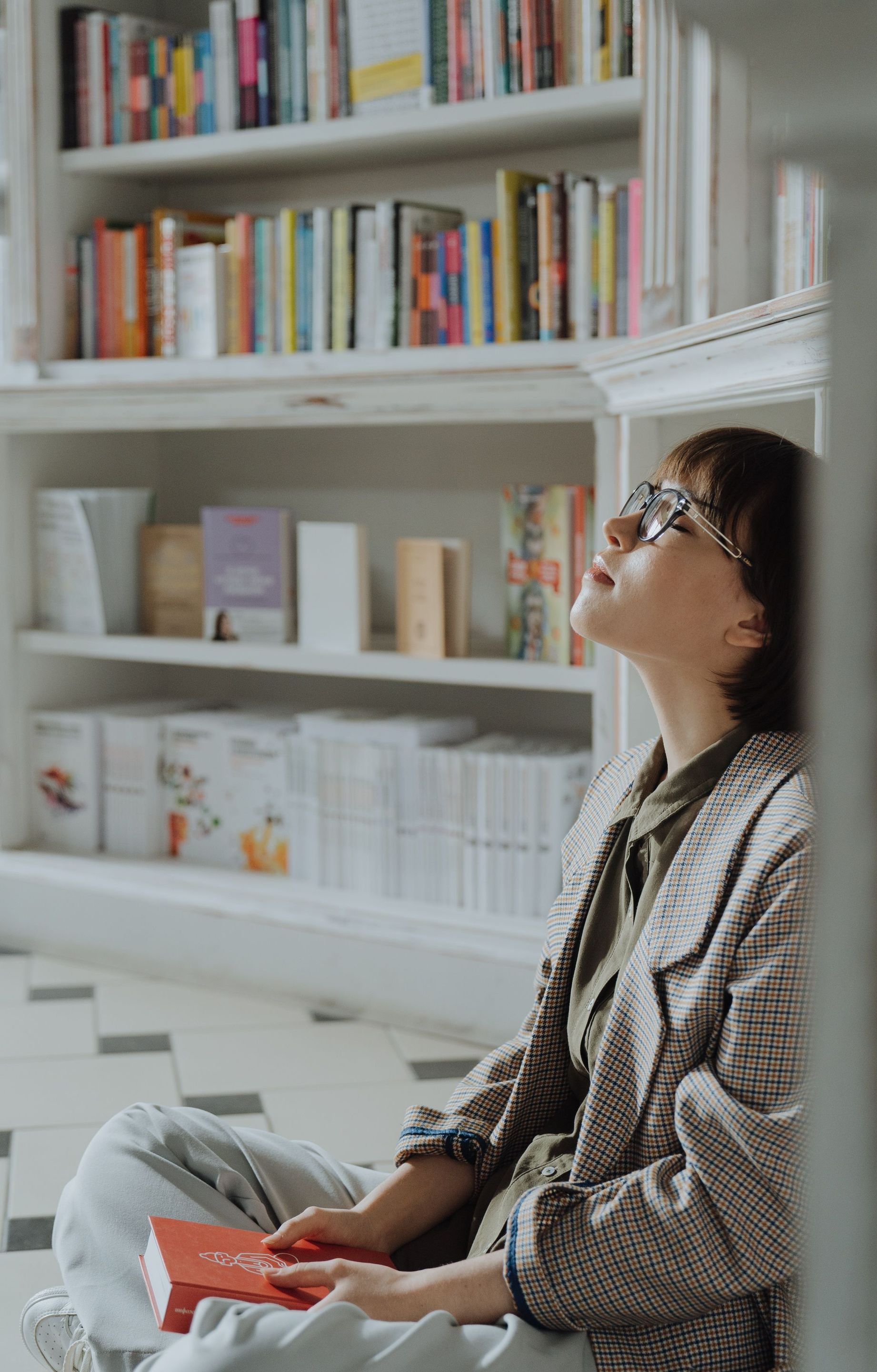 Woman sitting in a bright library, reading a red book and looking up at shelves of books