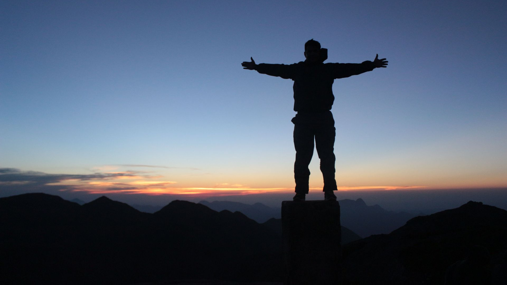 Silhouette of a person standing with arms outstretched on a peak at sunset over dark mountains
