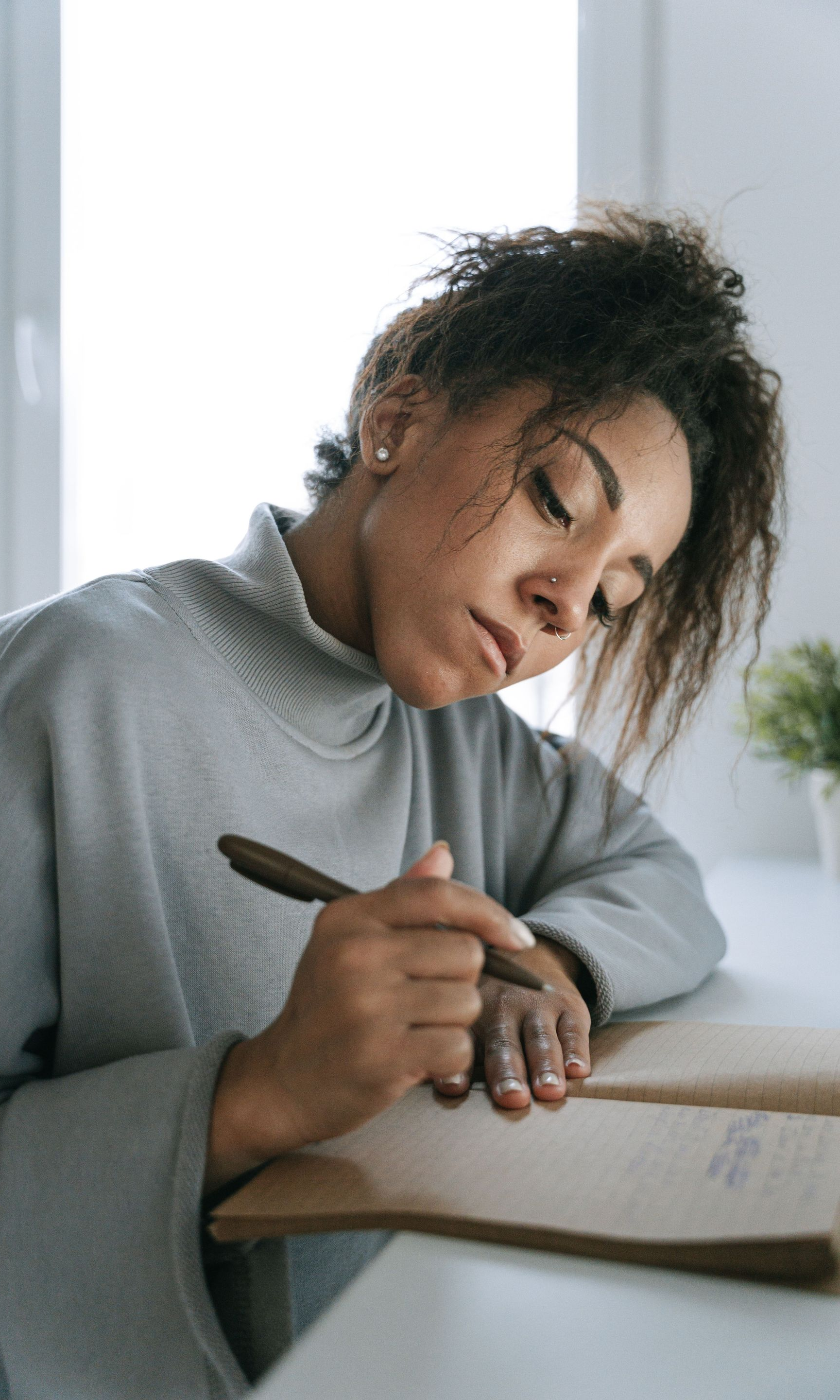 Person writing in a notebook at a desk, wearing a gray sweater, with a focused expression.