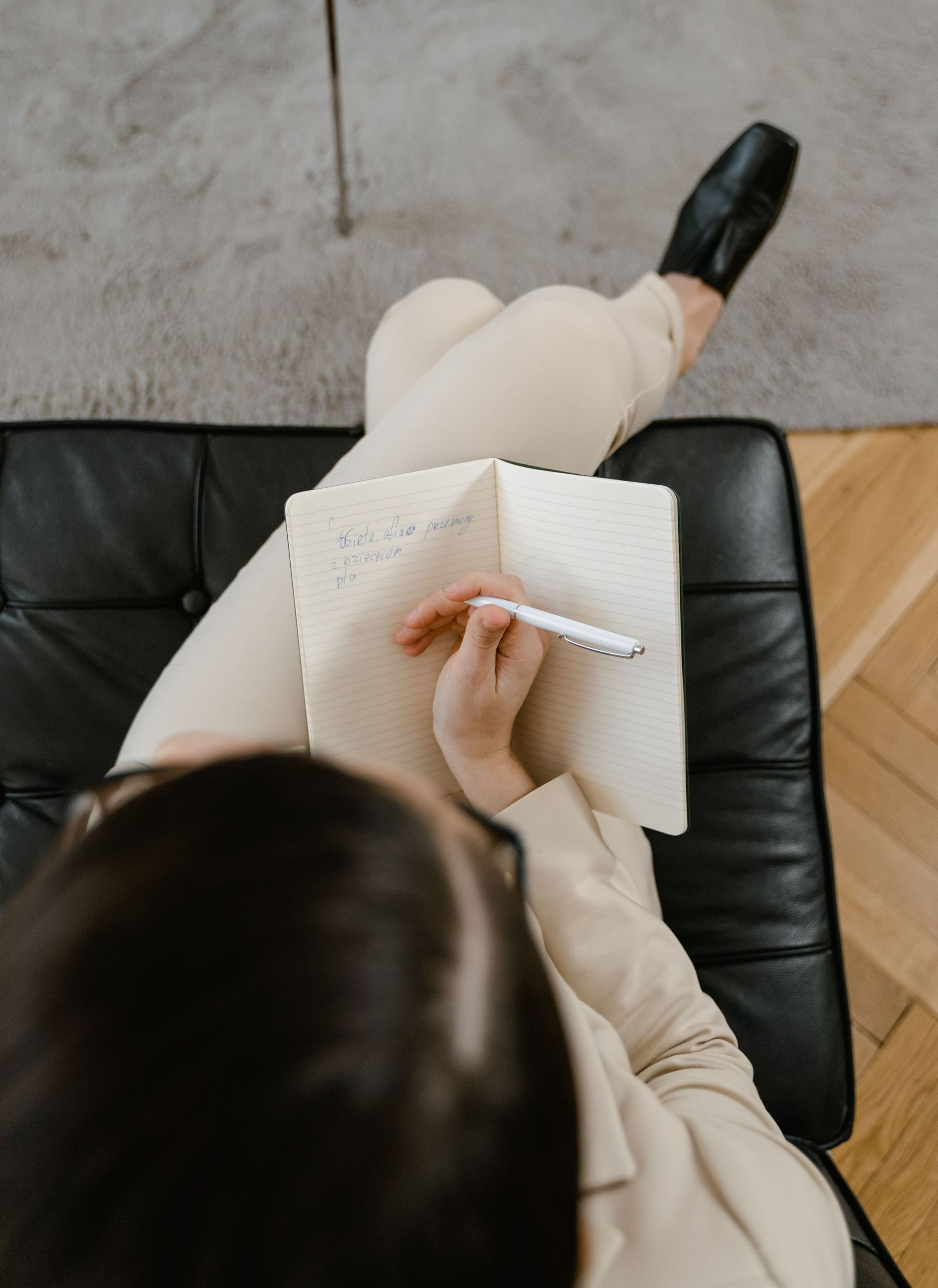 Person writing in a notebook while seated on a black chair, viewed from above.