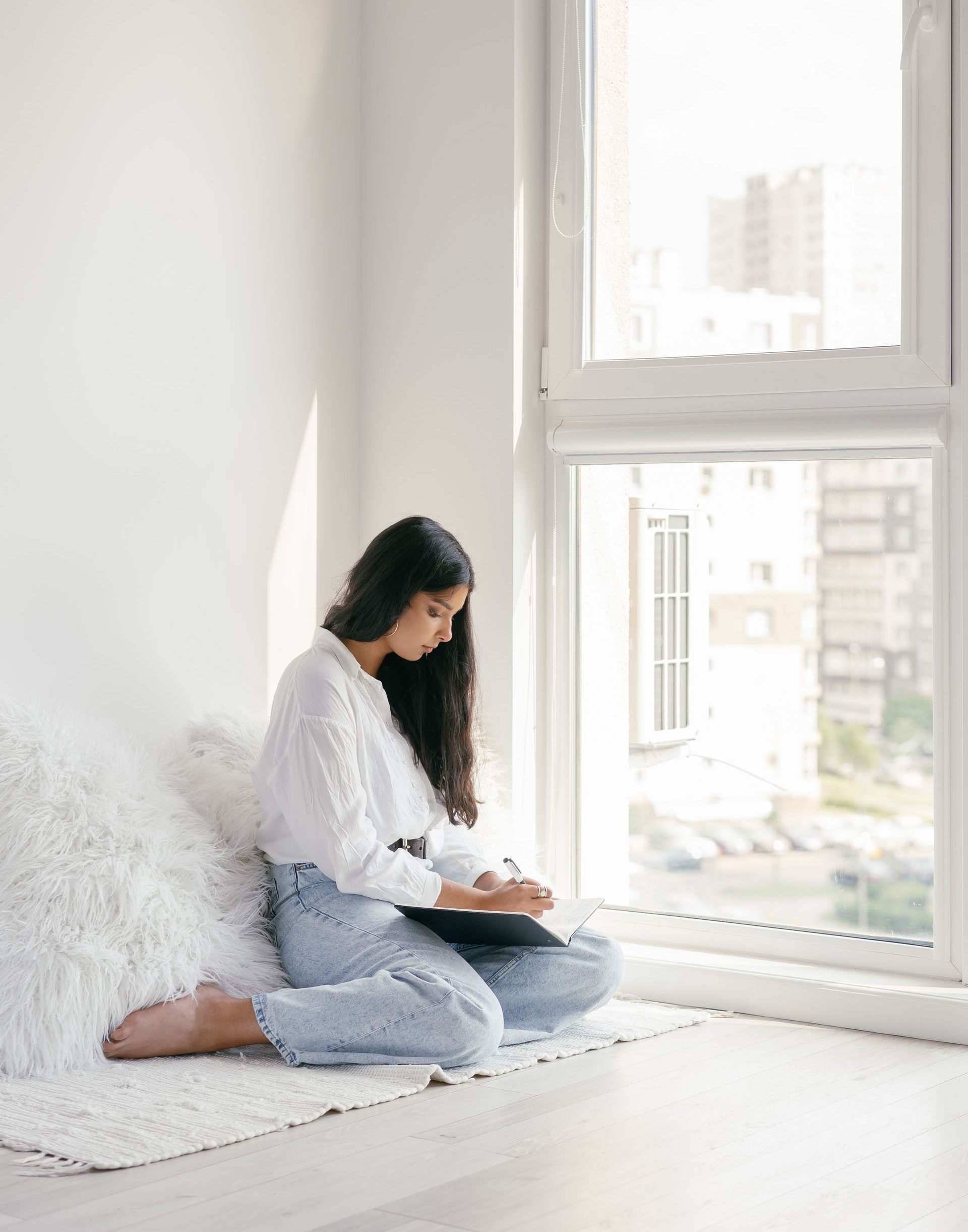 Woman sitting by a bright window, writing in a notebook on a white rug.
