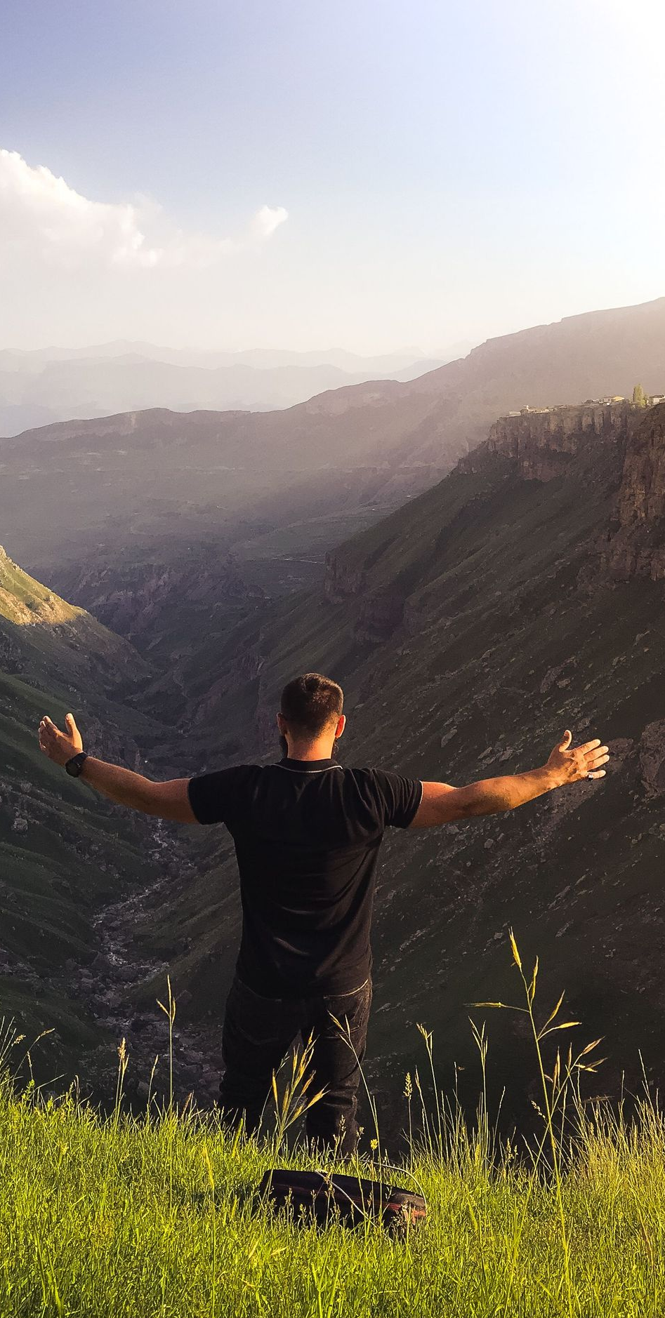 Person standing on a grassy mountain ridge with arms outstretched, overlooking a wide valley at sunset