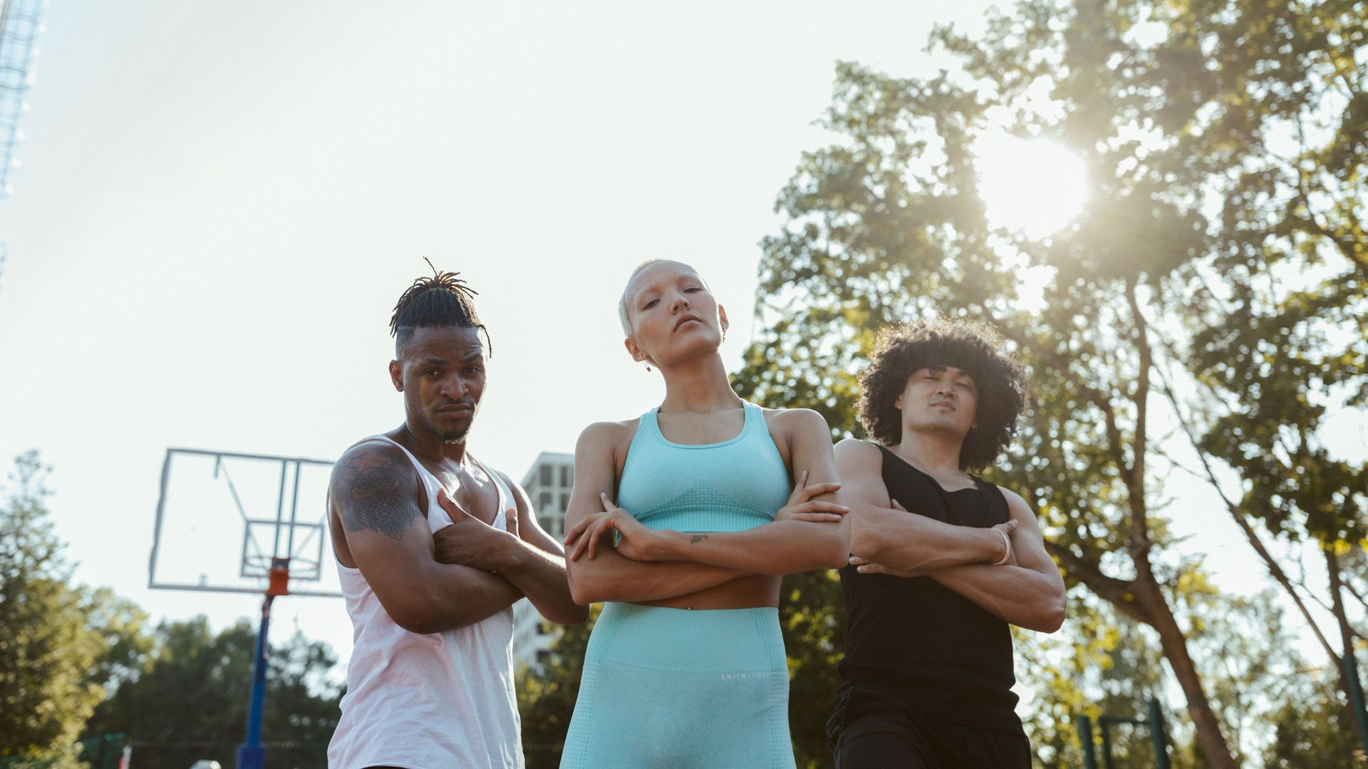 Three athletes stand with crossed arms on an outdoor basketball court in bright sunlight.