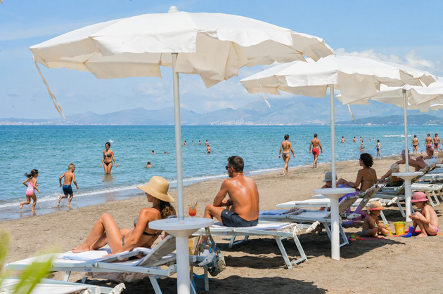 Spiaggia soleggiata con ombrelloni bianchi, lettini e persone che si rilassano in riva al mare.