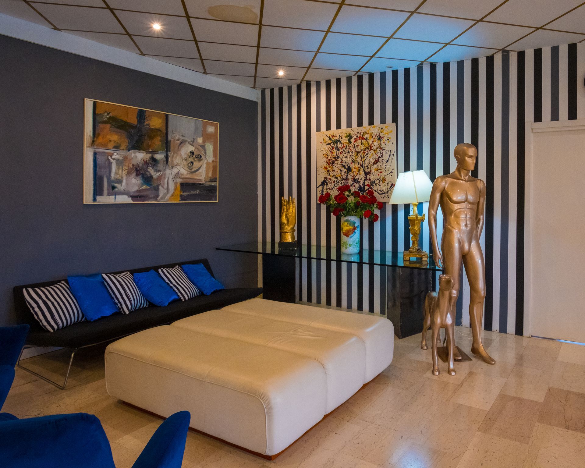 Soggiorno moderno con divani in velluto blu, pouf bianco, parete decorativa a righe e manichino dorato.