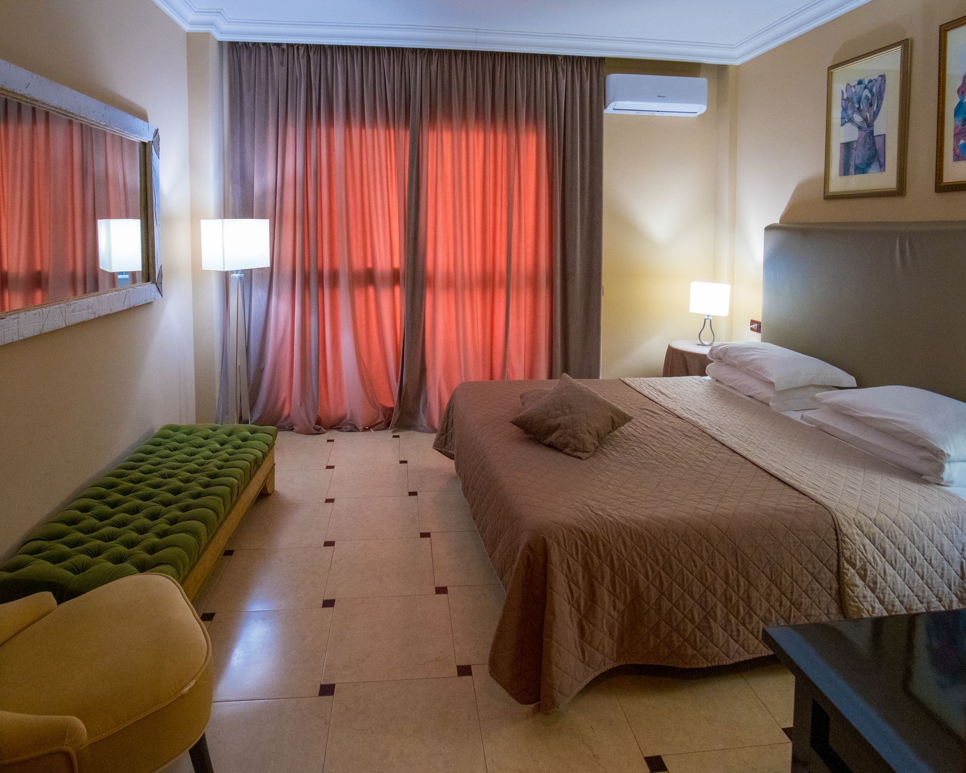 Camera d'albergo ben arredata con letto, tende alle finestre, poltrona e lampada da comodino.
