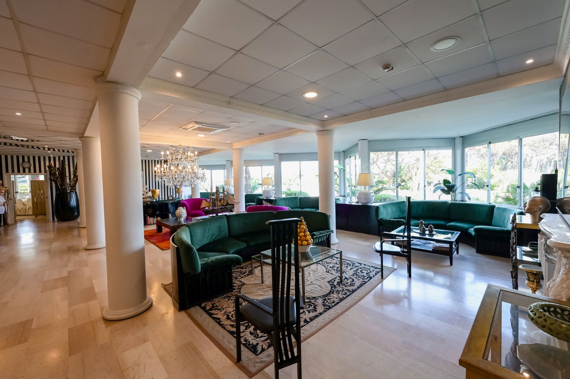 Luminosa hall dell'hotel con divani verdi, lampadari e ampie finestre che si affacciano sul verde.