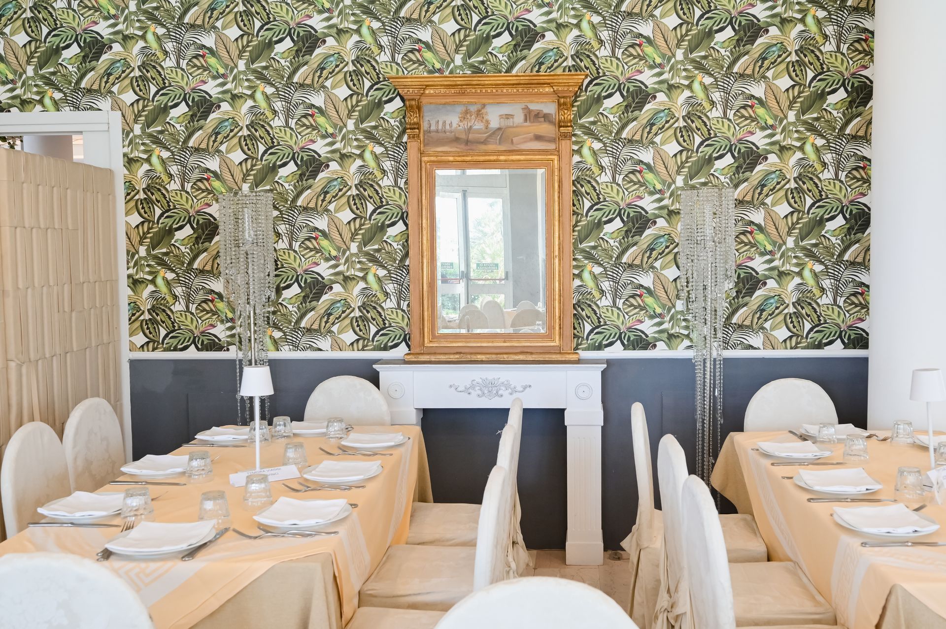 Elegante sala da pranzo del ristorante con carta da parati floreale, specchio e tavoli apparecchiati con cura con sedie bianche.