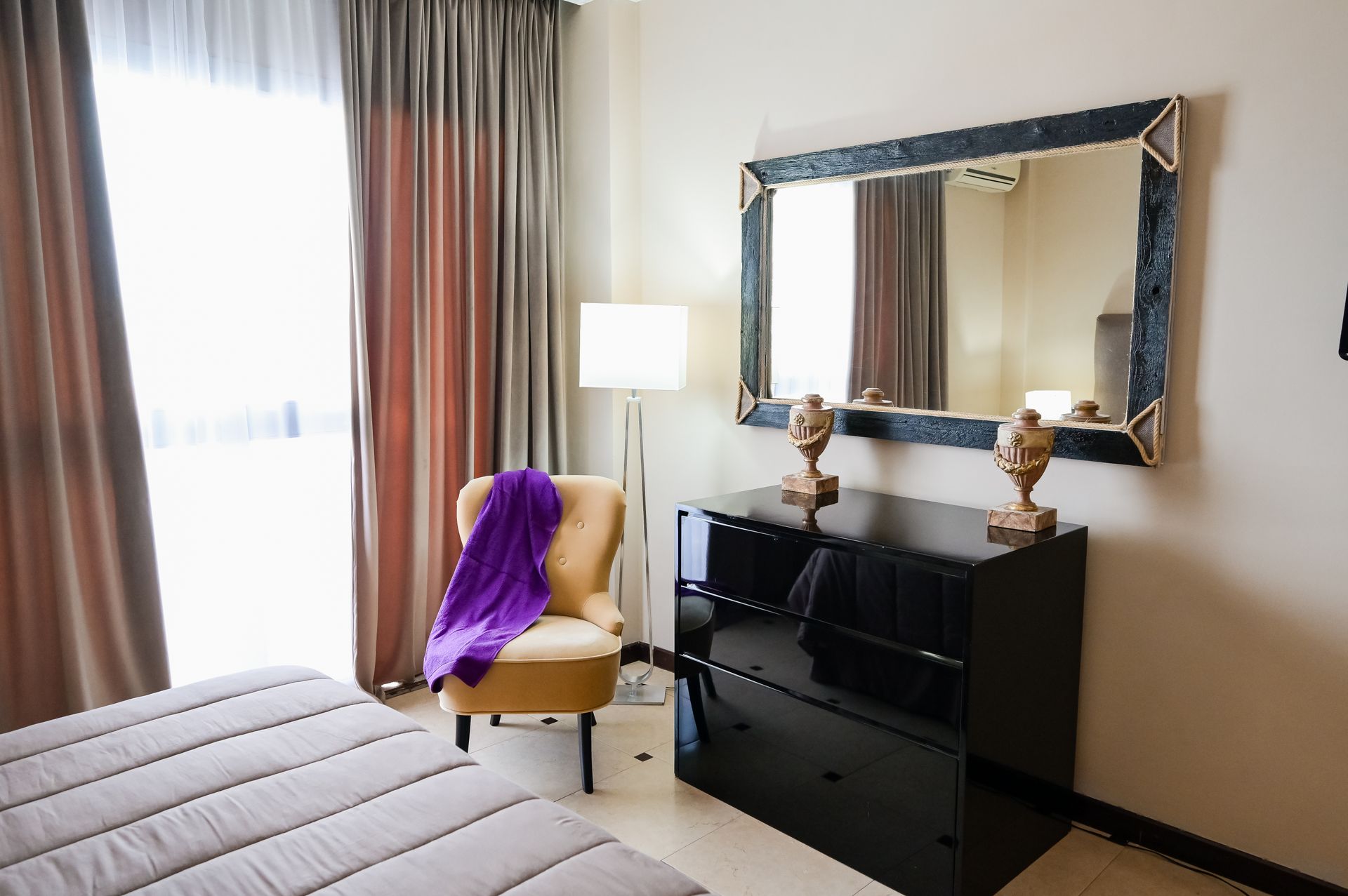 Camera da letto moderna con letto, sedia drappeggiata di viola, lampada e comò nero con un grande specchio.