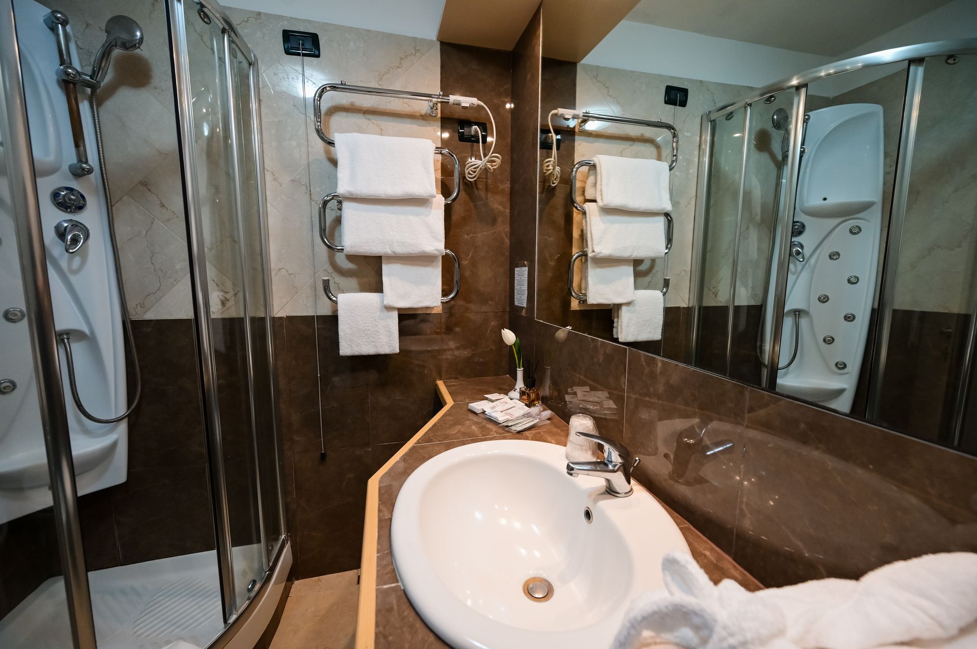 Bagno moderno con lavabo, specchi, doccia in vetro e asciugamani bianchi su pareti rivestite di piastrelle marroni.