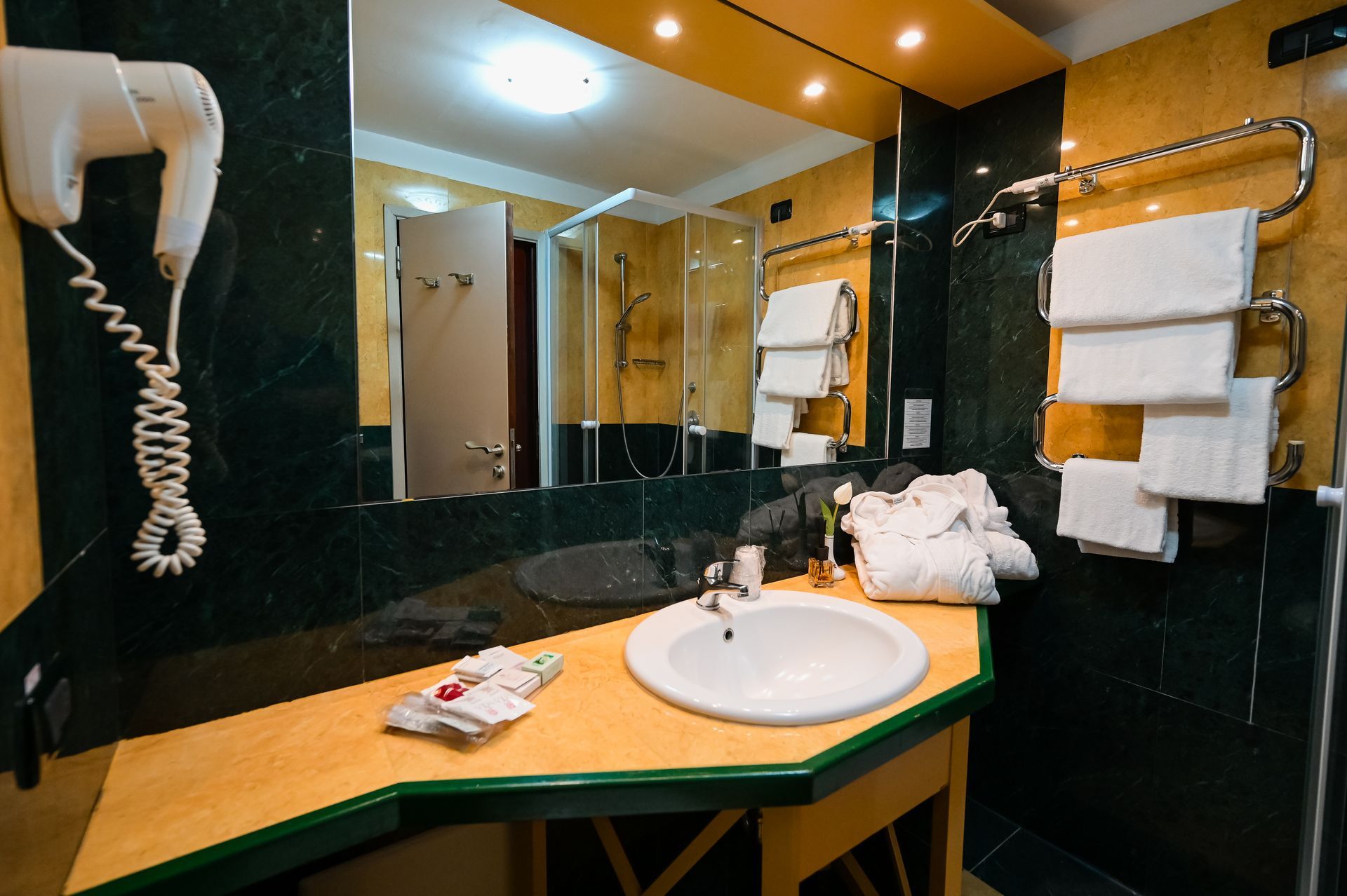 Lavabo del bagno con specchio, asciugacapelli a parete e asciugamani in un ambiente interno in stile hotel con luci calde.
