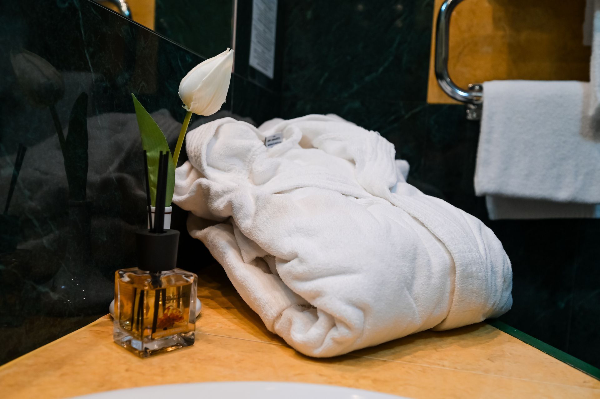 Asciugamani bianchi piegati su un ripiano accanto a un diffusore a bastoncini e un fiore bianco nella zona del lavabo del bagno.