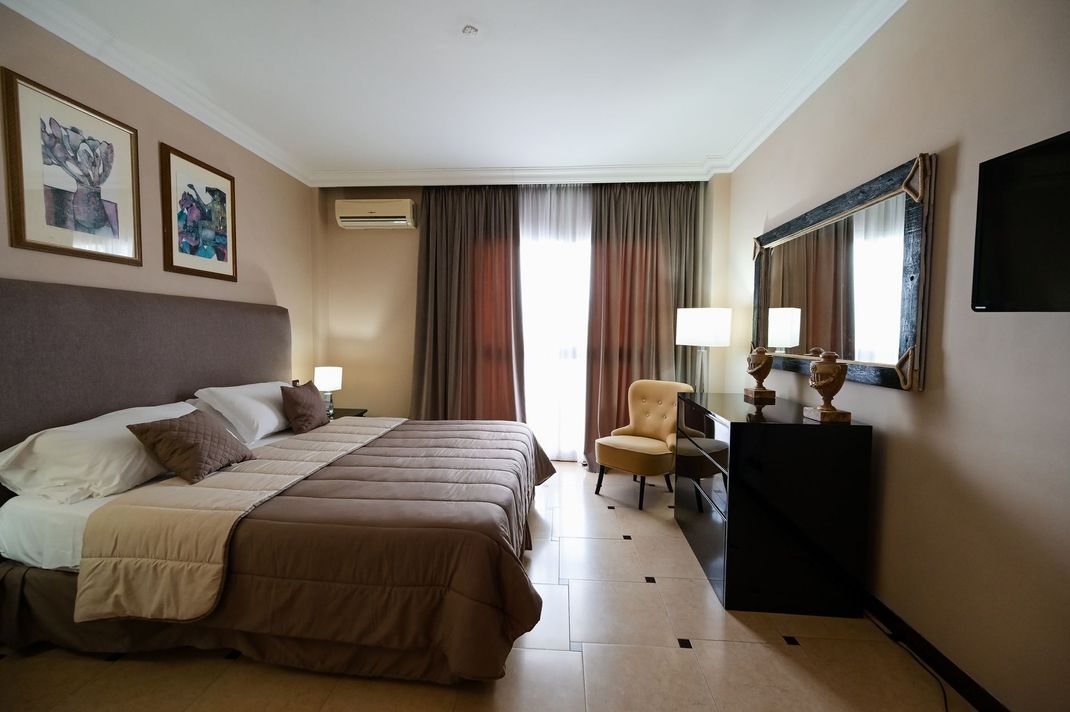 Camera d'albergo con letto, sedia, lampade da comodino, quadri alle pareti e finestra con tende.