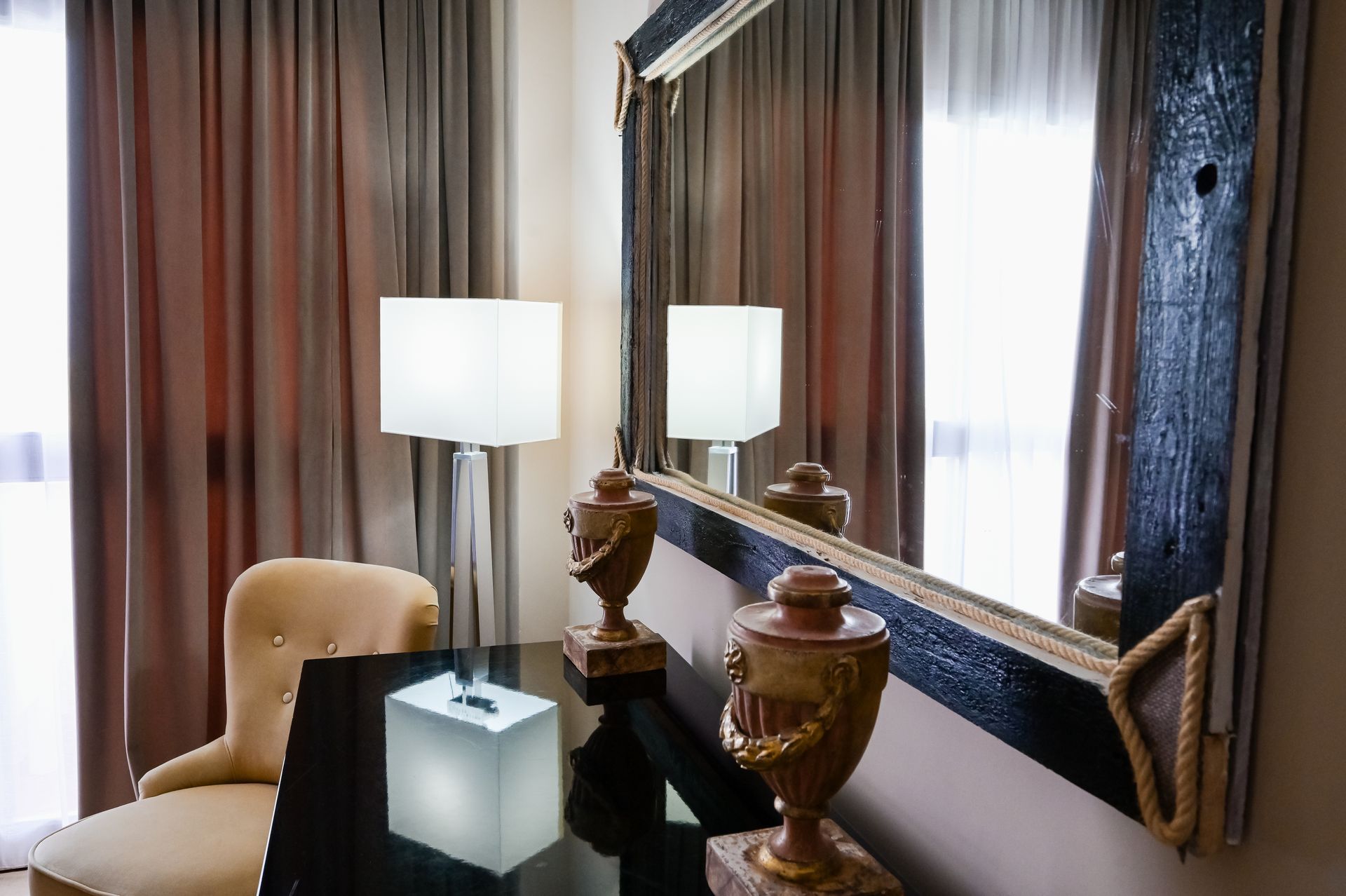 Elegante camera d'albergo con scrivania, sedia beige, lampada bianca e grande specchio accanto a tende marroni.