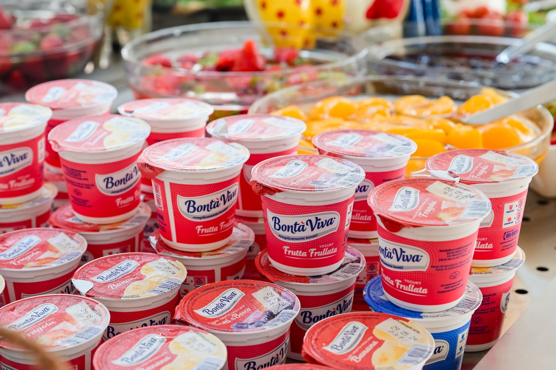 File di vasetti di yogurt rossi e bianchi su un banco del mercato, con frutta sullo sfondo.