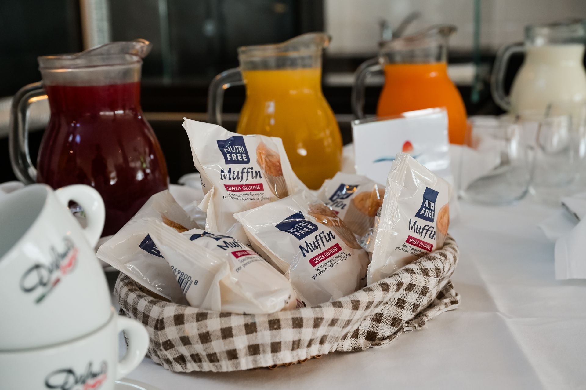 Un cesto di pacchetti di marmellata assortiti su un tavolo da colazione, con caraffe e tazze di succo sullo sfondo.