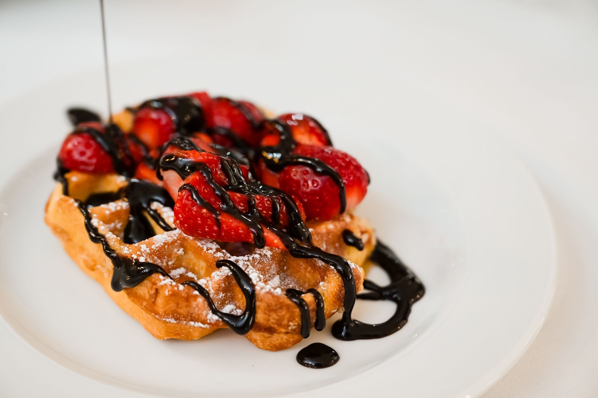 Waffle guarnito con fragole, glassa al cioccolato e sciroppo su un piatto bianco.
