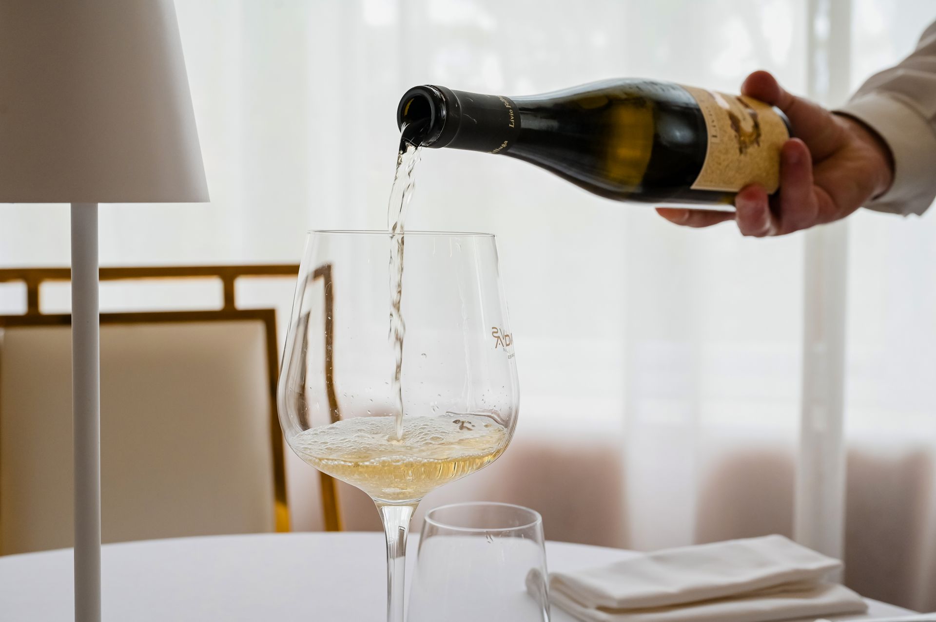 Versare a mano del vino bianco in un bicchiere accanto a una lampada da tavolo e a un tovagliolo piegato.